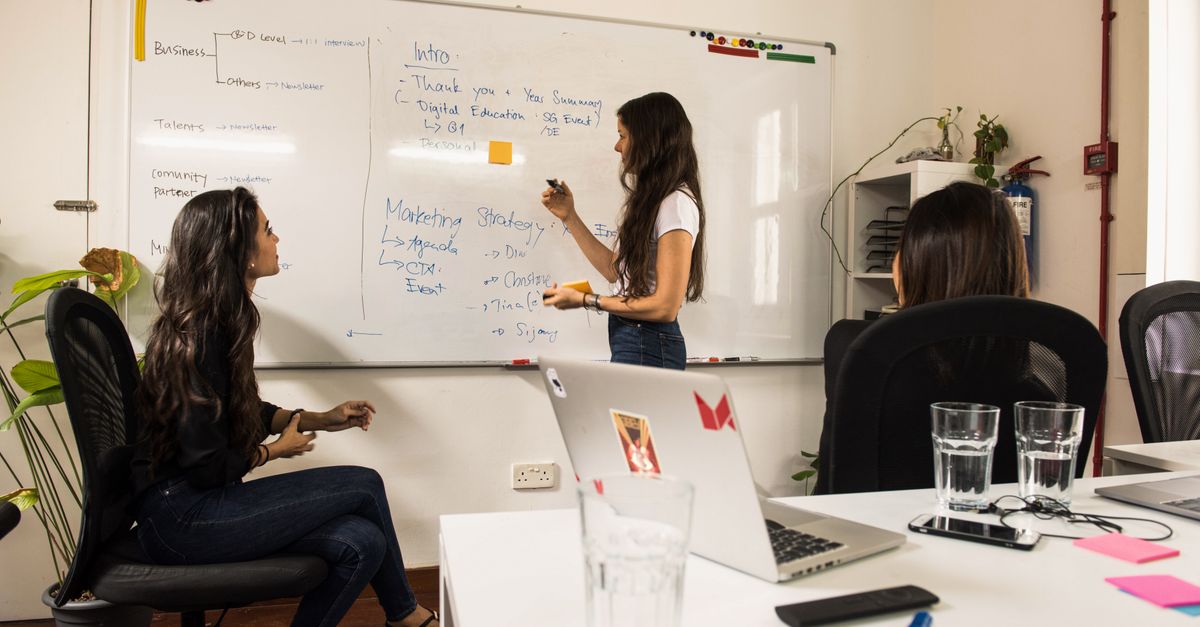 Business women discussing strategy at a whiteboard in a modern office setting.