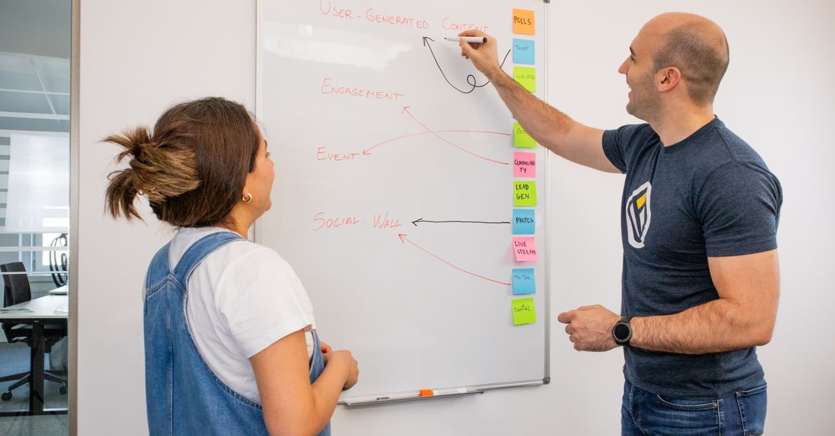 Two colleagues brainstorm marketing strategies on a whiteboard in a modern office setting.